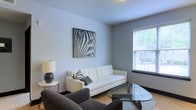 Located at 205 S Front St in Columbus, Ohio, this single-floor space offers a clean, contemporary aesthetic that prioritizes comfort and modern design. The living area serves as the centerpiece of the unit, showcasing neutral gray walls that create a calm, welcoming ambiance. A sleek white tufted sofa pairs effortlessly with a charcoal accent chair, anchored by a geometric glass coffee table that adds a touch of sophistication. A bold zebra-print wall canvas introduces a striking artistic focal point, giving the room a curated, gallery-like quality. Natural light filters in through a window fitted with horizontal blinds, brightening the space and providing a connection to the outdoors. Light hardwood-style flooring runs throughout, complementing the neutral palette and contributing to an open, airy feel. A stylish table lamp on a side table rounds out the living area with warm, ambient lighting. The overall design is cohesive and thoughtfully appointed, balancing minimalist sensibility with inviting warmth. Situated in the vibrant South Front Street corridor, this space enjoys a prime Columbus location close to dining, entertainment, and the scenic Scioto River waterfront.