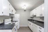 This single-floor unit offers a practical and functional layout across several well-defined spaces. The galley-style kitchen is outfitted with white cabinetry featuring black hardware, dark granite-look countertops, and white appliances including an electric range, refrigerator, and dishwasher. Light wood-look flooring and a ceiling light fixture complete the kitchen's clean, utilitarian aesthetic.

The open living area extends from the kitchen, finished with the same wood-look flooring and neutral gray walls. A sliding glass door with vertical blinds provides access to an outdoor patio, allowing natural light to fill the space. Adjacent to the living area, a foyer features warm wood-toned doors and a chandelier overhead.

The bedrooms are carpeted in neutral tones and each feature a window with wood-framed blinds, offering natural light and outdoor views. Textured ceilings are consistent throughout the unit.

Two bathrooms serve the space, both finished with wood-look flooring and white vanities topped with dark countertops. One bathroom includes a tub and shower combination with a sliding glass enclosure, while the other features a vanity with a framed mirror. Both bathrooms maintain a clean, straightforward design.