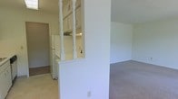 This single-floor residential space offers a practical and functional layout designed to maximize the use of its interior. A defining architectural feature is the half-wall partition adorned with white decorative spindle columns, which elegantly divides the kitchen from the living area while maintaining an open, connected feel throughout the space. The kitchen is equipped with white appliances, including a full-size refrigerator, and features light-toned cabinetry that provides ample storage. The flooring transitions from tile in the kitchen area to carpet in the living space, offering a clear visual distinction between the two zones. The living area is bathed in a neutral, light color palette with white walls that contribute to a bright and airy atmosphere. The space also includes a doorway visible in the background, suggesting additional rooms or storage beyond the main living area. Overall, the layout is straightforward and well-organized, making it a highly functional residential space suited for everyday living.