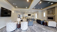 This community amenity space is designed to offer residents a comfortable and functional gathering environment. The interior lounge area features a sophisticated design highlighted by layered tray ceilings with recessed lighting, creating a refined architectural focal point overhead. The space is furnished with a mix of patterned accent chairs, plush sofas, and coordinating side tables, all arranged to encourage both relaxed conversation and casual socializing. Patterned carpeting in neutral tones ties the seating areas together cohesively.

Adjacent to the lounge, a kitchenette and bar-height counter area provides residents with convenient access to appliances, including a refrigerator and microwave, making the space well-suited for social gatherings and events. A wall-mounted television adds an entertainment element to the setting.

The outdoor area extends the amenities further with a generously sized swimming pool surrounded by a broad concrete sundeck. Rows of lounge chairs and patio umbrellas offer ample space for relaxation, while a pergola structure provides a shaded retreat. The pool area is enclosed by a perimeter fence, with the multi-story residential building serving as a backdrop, reinforcing the sense of a cohesive, well-planned community environment.
