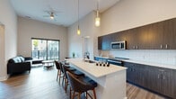 This single-floor residence showcases a contemporary design with soaring ceilings and an open, airy layout that seamlessly connects the main living spaces. The kitchen is a standout feature, equipped with dark espresso cabinetry extending to the upper reaches of the tall walls, a white subway tile backsplash, and a full suite of stainless steel appliances. A large central island with a crisp white countertop and integrated sink provides generous prep space and seating for multiple bar stools, making it ideal for both cooking and entertaining. Pendant lighting and a ceiling fan enhance the ambiance of the open-concept kitchen and living area, which flows directly to a private balcony through sliding glass doors, inviting abundant natural light and outdoor views.

The living area features smooth hardwood-style flooring, while the bedrooms transition to plush carpet. Each bedroom offers large black-framed windows with outdoor views, and tall ceilings contribute to a spacious feel. The primary bathroom is elegantly appointed with a long white vanity, a backlit mirror, and tile flooring. A dedicated walk-in closet with wire shelving provides ample storage. A separate in-unit laundry room with cabinetry and wood-style flooring adds practical convenience throughout.