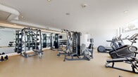 Located at 10401 Washington Blvd in Culver City, this professional-grade fitness center offers a comprehensive workout environment across a single, well-organized floor. The space features a modern, clean aesthetic with crisp white walls, neutral rubberized flooring, and a combination of recessed and ambient lighting that creates a bright, energizing atmosphere. Soaring ceilings in the central corridor are complemented by large floor-to-ceiling windows that invite abundant natural light and offer views of the surrounding landscape, enhancing the open and airy feel of the space. The strength training area is well-appointed with a multi-station cable machine, a full rack of dumbbells spanning a wide weight range, adjustable benches, and additional free weight equipment — all positioned alongside expansive wall mirrors that maximize the sense of space. The cardio zone features multiple treadmills, an elliptical, a stationary bike, and a rowing machine, lined along the windowed corridor for an engaging workout experience. A dedicated area houses a Pilates reformer and open floor space suitable for stretching or group training, with a wall-mounted flat-screen TV for entertainment. The layout flows intuitively from zone to zone, making this fitness center a fully functional and thoughtfully designed facility.