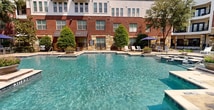This expansive outdoor amenity space is the centerpiece of a well-appointed multi-story apartment community, showcasing a classic blend of brick and natural stone architecture that frames the area with a distinctive, upscale aesthetic. The highlight of the space is a large, resort-style swimming pool with crystal-clear blue water, complemented by a shallow wading area featuring decorative stepping stones and an adjacent spa or splash zone. Surrounding the pool is a generously sized sun deck furnished with rows of white lounge chairs and shaded seating areas complete with patio umbrellas, ideal for relaxation outdoors. The courtyard areas are thoughtfully landscaped with ornamental grasses, agave plants, flowering shrubs, river rock accents, and mature shade trees that provide natural canopy throughout. Herringbone-patterned paver walkways connect the various zones of the outdoor space, adding a refined, cohesive design element. Private balconies and ground-floor patios with black iron railings overlook the amenity area from the surrounding building. Proper safety signage is posted at the pool entrance, and landscape lighting is integrated throughout the grounds. The overall ambiance is that of a private, resort-like retreat designed for both active recreation and quiet outdoor enjoyment.