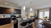 This single-floor residence located in Little Elm, Texas, offers a thoughtfully designed open-concept layout that seamlessly connects the kitchen, dining, and living areas. The kitchen is a standout feature, showcasing rich dark-stained cabinetry, granite countertops with subtle veining, and a full suite of stainless steel appliances, including a range, microwave, and refrigerator. A white subway tile backsplash adds a clean, contemporary contrast to the darker cabinetry. The central kitchen island provides generous counter space and doubles as a casual dining area, accommodating multiple bar-height seats beneath elegant pendant lighting with gold-toned accents. Track lighting overhead ensures the cooking area is well-illuminated throughout the day.

Wood-look flooring runs through the main living areas, lending warmth and cohesion to the space. The bedroom features plush carpeting, a ceiling fan, and a large window dressed with layered drapes, allowing for ample natural light. A walk-in closet with built-in shelving connects directly to the bathroom, which includes a soaking tub with a tiled surround and a shower combination. The overall ambiance of the home is modern and inviting, with neutral tones and stylish details creating a comfortable and functional living environment.