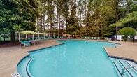 This outdoor pool facility in Woodstock, Georgia offers a well-appointed recreational retreat set against a backdrop of towering Georgia pines and mature landscaping. The centerpiece is a generously sized freeform swimming pool with a 5-foot depth, featuring vivid turquoise water and clean concrete decking that wraps around the perimeter, providing ample space for lounging and movement.

Surrounding the pool, a large collection of teal lounge chairs are arranged along the deck, offering plenty of seating for sunbathers. A white pergola structure provides a shaded zone along one side of the deck, lined with additional chaise lounges for guests seeking relief from the sun. Umbrella-covered tables and chairs are also available near the clubhouse for casual outdoor dining or relaxation.

A two-story clubhouse anchors the pool area, constructed with a stone facade on the lower level and white-painted wood detailing on the upper level, which features an open balcony with decorative railings. The lower covered porch area provides additional sheltered seating and houses facility signage and safety equipment.

The entire pool area is enclosed by a black wrought-iron fence, ensuring security and privacy. The lush tree canopy surrounding the space creates a serene, park-like atmosphere, making this pool area a standout community amenity in Woodstock, GA.