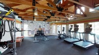 This fitness center is housed in a striking single-story space defined by its dramatic exposed wood beam and truss ceiling, which rises to impressive heights and lends the room a warm, open, lodge-like character. Industrial-style track lighting and metal bracket connectors complement the natural wood tones overhead, while large windows and glass doors along multiple walls flood the interior with abundant natural light and provide views of the surrounding outdoor greenery.

The workout floor is covered in durable dark rubber flooring and organized to accommodate a variety of exercise routines. The cardio zone features multiple treadmills, elliptical machines, and stationary bikes, many equipped with individual screen monitors. A dedicated strength training area includes a Life Fitness functional trainer cable machine with battle ropes and resistance attachments, as well as a full dumbbell rack with an adjustable bench. Additional benches and storage seating provide convenient rest areas between sets.

A bold horizontal stripe mural in red, orange, and yellow wraps the upper walls, adding a dynamic, energetic ambiance throughout the space. A large graphic wall mural featuring athletic figures anchors one end of the room. Practical amenities include a water fountain station, sanitation dispensers, and restroom access. The fitness center is thoughtfully designed to support a comprehensive workout experience in a visually engaging environment.