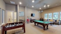 This single-floor amenity space is thoughtfully designed to deliver a comprehensive entertainment experience, divided into two distinct areas: a lively game room and a private screening room. The game room is generously sized, featuring neutral-toned walls, recessed lighting, and soft carpet flooring throughout. It is equipped with a full-size pool table illuminated by stylish pendant lighting, a foosball table, and a wall-mounted flat-screen TV. A popcorn machine adds a festive touch, while a water fountain, a lounge sofa with views of the surrounding greenery through large windows, and a bar-height table with stools round out the social atmosphere. Large windows along one wall invite abundant natural light and offer pleasant outdoor views of the landscaped surroundings. The screening room is a dedicated, intimate theater space furnished with multiple rows of black leather recliner seats equipped with cup holders, offering a true cinema-style experience. A large projection screen anchors the room, while classic Hollywood-inspired wall art adds a touch of personality to the space. Together, these two areas create a versatile and inviting venue well-suited for casual gatherings, movie nights, and recreational activities.