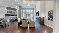 This elegantly designed single-floor community lounge offers a refined and welcoming atmosphere defined by soaring ceilings and rich, dark hardwood flooring throughout. The main gathering area is anchored by a sleek, contemporary fireplace with a mounted flat-screen television above, surrounded by multiple thoughtfully arranged seating groupings that include plush sofas, curved accent chairs, and stylish side tables. A striking sputnik-style chandelier draws the eye upward, complementing the recessed lighting that illuminates the open layout. Floor-to-ceiling windows and glass-panel doors flood the space with abundant natural light while offering views of the surrounding exterior courtyard, creating a bright and airy ambiance. Textured accent columns add architectural depth and visual interest throughout the room. A separate dining area features round glass-top tables paired with upholstered chairs in a soft blue-grey palette, set beneath equally lofty ceilings. The space also includes a fully equipped kitchen outfitted with dark wood cabinetry, white subway tile backsplash, light tile flooring, and stainless steel appliances including a side-by-side refrigerator and dishwasher. Together, these areas create a cohesive, multi-functional environment ideal for both relaxation and social gatherings.