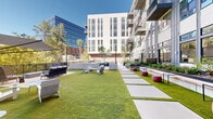 Located at 600 Saint Albans Drive in Raleigh, NC, this expansive outdoor amenity space is a masterfully landscaped courtyard set within a modern multi-story residential community. The design seamlessly blends open green lawn areas with structured hardscape zones, creating a dynamic environment suited for both relaxation and social gatherings.

A central turfed lawn is anchored by large concrete stepping stone pathways that guide visitors through the space. Along the perimeter, raised dark metal planters are filled with ornamental grasses, shrubs, and young trees, adding layered greenery and natural texture throughout. Slatted wood benches integrated into the planter edges provide casual seating in close proximity to nature.

Multiple distinct seating zones are distributed across the courtyard, ranging from lounge chairs and Adirondack-style furniture to modular ottomans and bar-height tables. A striking green-framed outdoor bar structure serves as a focal point in one section, complemented by bistro string lights overhead that add ambiance during evening hours.

A raised terrace along one edge offers bar seating with views of the surrounding tree canopy and neighboring skyline. The contemporary architectural backdrop, featuring a mix of glass curtain walls, horizontal siding, and brick accents, frames the courtyard and connects interior spaces to the outdoors through large glazed entryways.