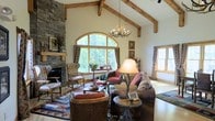 This single-floor space exudes rustic elegance through a thoughtful combination of natural materials and warm, inviting design. The living area serves as the heart of the home, commanding attention with its dramatic vaulted ceiling adorned with exposed timber beams that add both architectural interest and a sense of grandeur. A striking floor-to-ceiling stacked stone fireplace anchors one wall, providing a focal point that enhances the cabin-inspired character of the space. An antler chandelier hangs overhead, complementing the nature-themed aesthetic with an authentic rustic touch. Natural light pours in through a large arched window framed in warm wood, offering picturesque views of the surrounding greenery. Additional windows reinforce the connection to the outdoors and keep the interior bright and airy. The living area is furnished with plush, earth-toned seating, layered rugs with Southwestern-inspired patterns, and organic accent pieces that harmonize with the overall design language. Every detail — from the textured stone to the handcrafted wood elements — contributes to an atmosphere that is both cozy and sophisticated, making this space feel like a welcoming woodland sanctuary.