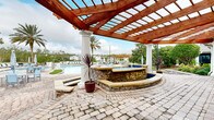 Located at 265 Shady Oaks Dr in Palm Coast, Florida, this outdoor community amenity space offers a resort-inspired experience designed for relaxation and recreation. The centerpiece is a generously sized swimming pool with a classic freeform shape, finished with blue tile accents and accessible via stainless steel entry rails. The pool is surrounded by an expansive paver deck lined with lounge chairs and umbrella-shaded seating areas, providing ample space for sunbathing and socializing.

A standout architectural feature is the bold wood pergola supported by elegant white classical columns, beneath which sits a raised stone-clad spa. The natural stone masonry and warm wood tones of the pergola create a striking contrast against the Florida sky, lending the area a sophisticated, upscale character. Decorative potted tropical plants and mature palm trees enhance the lush, resort-like ambiance throughout the grounds.

Adjacent seating clusters with tables and chairs offer additional gathering areas, while the surrounding landscaping—including manicured hedges, flowering shrubs, and a tree-lined perimeter—provides privacy and a natural backdrop. A charming clubhouse structure with a distinctive cupola is visible from the pool area, anchoring the space within a well-appointed residential community setting.