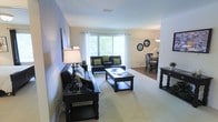 This single-floor residence offers a thoughtfully designed open-concept layout that seamlessly connects the living and dining areas, creating a cohesive and inviting atmosphere. The living area is anchored by plush wall-to-wall carpeting and features an arrangement of dark leather seating accented with green throw pillows, complemented by a substantial dark wood coffee table and console. A large picture window dressed with sheer white curtains floods the space with natural light and offers views of lush greenery outside, enhancing the bright and airy feel of the room. The dining area is positioned adjacent to the living space and is furnished with a classic dark wood dining set, coordinating seamlessly with the overall design palette. Decorative circular mirrors adorn the wall, adding depth and a stylish finishing touch to the shared space. A pendant light fixture overhead provides warm ambient lighting for the dining area. The neutral wall tones and light-colored ceilings create a clean, spacious backdrop that ties the entire living space together. Overall, the residence presents a balanced blend of comfort and classic elegance, making it an inviting and functional home environment.
