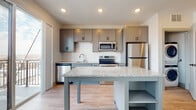 This well-appointed single-floor unit offers a thoughtfully designed layout with modern finishes throughout. The kitchen serves as the centerpiece of the space, showcasing gray shaker-style cabinetry with brushed hardware, a crisp white subway tile backsplash, and light granite countertops. A substantial kitchen island with built-in open shelving provides additional prep space and storage. The full suite of stainless steel appliances includes a refrigerator, electric range, over-the-range microwave, and dishwasher, delivering both style and functionality. Light wood-tone flooring runs throughout, lending a warm, cohesive feel to the interior. Recessed lighting illuminates the kitchen and living areas with a clean, contemporary look. The bathroom features white shaker-style cabinetry, a single-sink vanity with a wide mirror, and a tub-shower combination, all finished with the same light wood flooring for consistency. A sliding glass door leads to a private balcony, flooding the adjacent dining area with natural light and offering open outdoor views. A window with horizontal blinds adds additional brightness to the space. The overall design is clean, modern, and functional, making excellent use of the available layout.