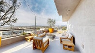 Located at 7650 Van Nuys Blvd in the Van Nuys neighborhood of Los Angeles, this rooftop terrace offers a well-appointed outdoor retreat with sweeping panoramic views of the surrounding cityscape and the San Gabriel Mountains beyond. The open-air deck features a clean, contemporary design characterized by smooth light-gray flooring, stucco-finished walls, and recessed ceiling lighting under a partially covered section that provides shade without sacrificing the open-sky ambiance.

The terrace is furnished with multiple teak-framed seating arrangements, including sofas and armchairs fitted with plush white cushions and colorful accent pillows, creating comfortable conversation areas. A large bowl-shaped fire pit serves as a central focal point on the open portion of the deck, surrounded by the seating groupings. A striking oversized yellow planter housing a small ornamental tree adds a bold pop of color and a touch of greenery to the space.

String lights are strung overhead across the terrace, lending a warm, inviting atmosphere during evening hours. Sun loungers positioned near the railing offer an ideal vantage point to take in the expansive views of Van Nuys Boulevard below and the mountain ridgeline in the distance. The rooftop's elevated position provides an impressive sense of openness, making it a versatile space suited for both relaxed lounging and social gatherings.