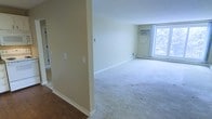 This single-floor space presents a straightforward and functional layout designed for comfortable everyday living. The main living area is finished with soft, light-colored wall-to-wall carpeting that adds warmth underfoot, while the surrounding walls are painted in a neutral, pale yellow-green tone that keeps the interior feeling bright and open. A large window along the far wall allows a generous amount of natural light to fill the room, with views of surrounding greenery visible from within, creating a pleasant connection to the outdoors. A wall-mounted air conditioning unit sits beneath the window, ensuring climate control throughout warmer months. A baseboard heating unit is also present, providing supplemental warmth during cooler seasons. The entry area transitions from hard flooring — likely wood or laminate — into the carpeted living space, offering a clear delineation between zones. The clean, uncluttered walls with standard white baseboards throughout give the space a tidy and well-maintained appearance. Overall, the space offers a calm, neutral canvas that is well-suited for a variety of furnishing styles and personal touches.