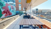 This space is part of a contemporary multi-story building and consists of a well-designed interior corridor paired with access to a striking rooftop-level outdoor terrace. The interior hallway features neutral white walls, carpeted flooring with a distinctive gray tile pattern accented by a bold red runner, and recessed overhead lighting. Wood-toned doors line the corridor, and convenient amenities such as vending machines are accessible along the way. The clean, functional design creates a bright and navigable passage throughout the building's floor.

The outdoor terrace is a standout feature of the space, offering an open-air setting elevated above the surrounding urban landscape. Furnished with sturdy wood-slatted dining tables and woven chairs arranged in a linear layout, the terrace comfortably accommodates multiple groups. Overhead infrared heaters and a retractable canopy provide year-round usability, while strands of globe string lights add a warm, inviting ambiance. A colorful large-scale mural adorns the adjacent brick wall, adding an artistic character to the setting. Open-railing perimeter fencing frames sweeping views of the city skyline, making this terrace an ideal space for gatherings or relaxation in an urban environment.