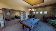 Located at 700 E Rodeo Rd in Casa Grande, Arizona, this single-story community clubhouse offers a well-appointed gathering space designed for comfort and recreation. The centerpiece of the facility is a spacious game room anchored by an ornate, carved-wood billiard table with a blue felt surface, suspended beneath an elegant wrought-iron chandelier. A decorative tray ceiling with warm golden tones adds architectural character to the room, while a row of high windows along one wall allows natural light to filter in. Patterned carpet and bistro-style seating areas along the perimeter create an inviting atmosphere for socializing. French doors with glass panes connect the game room to an adjacent area, enhancing the open flow of the space. The lounge area features plush, floral-upholstered sofas, a carved wood coffee table, and framed artwork on warm taupe walls, with a grand arched entryway leading to an outdoor courtyard. A compact kitchenette rounds out the amenities, equipped with a refrigerator, range, microwave, coffee station, and light wood cabinetry set against dark countertops and tile flooring—providing convenient refreshment options for all who gather here.