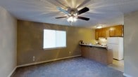 This single-floor space offers a comfortable and functional layout designed for everyday living. The main living area is finished with soft wall-to-wall carpeting underfoot and neutral-toned walls that create a warm, inviting atmosphere. A ceiling fan with an integrated light fixture is centrally mounted, providing both air circulation and ambient lighting. A window fitted with blinds allows natural light to filter into the room while maintaining privacy.

The living area flows seamlessly into the kitchen, which is separated by a raised breakfast bar — a practical and social feature that connects the two spaces without sacrificing definition. The kitchen is outfitted with warm wood-toned cabinetry, a white appliance suite, and under-cabinet lighting that adds a functional brightness to the cooking area. The breakfast bar provides additional counter space and can serve as a casual dining area.

The textured ceiling throughout adds a traditional residential character to the space. Overall, the layout is efficient and well-connected, making it easy to move between the living and kitchen areas. The neutral color palette and consistent flooring choices lend the space a cohesive and clean aesthetic, well-suited for a range of furnishing styles.