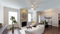 This well-appointed single-floor residence offers a warm and inviting atmosphere highlighted by thoughtful design details throughout. The living area serves as the heart of the home, centered around a classic tiled fireplace framed by a crisp white mantel and surround, adding both charm and a focal point to the room. Crown molding runs along the ceiling, lending an elegant architectural touch that complements the neutral, light-toned walls. Rich hardwood flooring extends across the living space, providing warmth and contrast to the soft, bright color palette. A ceiling fan with integrated lighting keeps the space comfortable and well-lit, while multiple windows dressed with white blinds allow abundant natural light to fill the room. The open layout connects the living area naturally to a dining space, visible in the background, making the floor plan feel expansive and functional. Recessed lighting adds additional illumination, and the overall ambiance is clean, airy, and modern with classic undertones — an ideal blend of comfort and style suited for everyday living and entertaining alike.