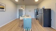 This contemporary single-floor unit offers a thoughtfully designed layout that balances functionality and modern style throughout. Light wood-tone flooring flows seamlessly across the entire space, complemented by a neutral gray-and-white color palette that creates a clean, cohesive aesthetic.

The open-concept kitchen is a standout feature, equipped with stainless steel appliances including a refrigerator, range, and built-in microwave. Sleek gray cabinetry pairs with a crisp white countertop island that doubles as a breakfast bar with seating, illuminated by modern pendant lighting above.

The living area is bright and airy, with floor-to-ceiling sliding glass doors opening onto a private balcony that frames expansive outdoor views, including a scenic mountain backdrop. Natural light fills the room throughout the day.

The bedroom is a calm, well-proportioned retreat with a window allowing in natural light. It connects directly to a private en-suite bathroom featuring a wide white vanity with ample storage, a large mirror, and a glass-enclosed shower.

A generously sized walk-in closet with built-in shelving and hanging rods provides excellent storage. In-unit stacked laundry is conveniently located nearby, adding practical day-to-day functionality to this well-appointed space.