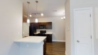 Located at 2030 Wildts Battery Blvd on Johns Island, South Carolina, this single-floor residence offers a well-designed layout with modern finishes throughout. The open-concept kitchen serves as the heart of the home, showcasing rich dark wood cabinetry that provides ample storage and a striking contrast against the light-toned walls. A spacious white countertop island anchors the kitchen, offering additional prep space and a natural gathering point. Black appliances, including a refrigerator, range, and over-the-range microwave, lend a cohesive, contemporary look to the culinary space. Overhead, a combination of track lighting and elegant pendant fixtures illuminates the kitchen with both functional and ambient light. A decorative chandelier is visible in an adjoining area, adding a refined touch to the overall aesthetic. The hardwood-style flooring runs throughout, delivering warmth and a sense of continuity across the living spaces. The neutral wall palette and clean architectural lines create a bright, welcoming ambiance that is both versatile and timeless. The single-floor layout ensures convenient, accessible living in a well-appointed setting on scenic Johns Island.