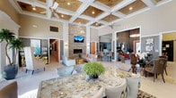 This single-story community clubhouse is designed with upscale finishes and a welcoming ambiance throughout. The centerpiece of the space is a expansive open-concept lounge and gathering area distinguished by soaring coffered ceilings with warm wood inlays, dual ceiling fans, and recessed lighting. A floor-to-ceiling stacked stone accent wall anchors the room, featuring a linear fireplace and wall-mounted television, while plush seating arrangements create an inviting atmosphere for relaxation or socializing.

Adjacent to the main lounge, a sunlit sitting area offers tall windows and French doors that open directly to an outdoor pool area, allowing natural light to flood the space and providing picturesque views of the surrounding community. This transitional area is furnished with high-back upholstered chairs and rustic wood-paneled accent walls, balancing elegance with comfort.

The kitchen area is thoughtfully appointed with slate-blue cabinetry, quartz countertops, a mosaic tile backsplash, stainless steel appliances including a full-size refrigerator and built-in microwave, and ample storage. A separate galley-style prep space mirrors these finishes with a utility sink and arched transom window.

A private office or concierge area features a sliding barn door, arched window, tiled floors, and workstation surfaces, completing this well-rounded and polished community facility.