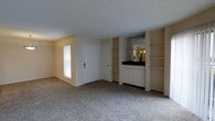 This single-floor residential space presents a practical and straightforward layout centered around an open living area. The room is finished with wall-to-wall carpeting in a neutral tone, contributing to a quiet and comfortable atmosphere. A textured ceiling adds subtle dimension overhead, while the light beige walls create a warm, cohesive backdrop throughout.

A standout feature of the space is the built-in shelving unit flanking a wet bar or kitchenette area along one wall. The built-ins include open shelves on both sides, ideal for display or storage, and a lower cabinet section with white cabinetry topped by a dark countertop. A mirrored panel above the counter reflects light and provides a sense of added depth to the room.

Natural light enters through a window fitted with horizontal blinds, brightening the space without overwhelming it. A white interior door provides access to an adjacent room or closet, maintaining a clean and uncluttered aesthetic. The overall design is functional and understated, with built-in storage solutions that maximize utility while keeping the living area open and accessible.