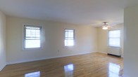 This single-floor residence offers a clean, well-maintained interior that combines classic design with comfortable, functional living. The main living area is a standout feature, showcasing polished hardwood floors that reflect natural light beautifully, creating a warm and inviting atmosphere throughout the space. Multiple double-hung windows line the walls, flooding the room with abundant daylight while offering views of the surrounding greenery outside. The neutral, cream-toned walls provide a versatile backdrop, complementing the natural wood tones of the flooring and lending the space a bright, airy feel. A classic ceiling fan with a light fixture is centrally positioned, ensuring both comfort and illumination. Clean white baseboards and trim add a crisp, finished look to the interior. The open layout of the living area allows for flexible furniture arrangements and a generous sense of space, making it well-suited for both relaxed everyday living and entertaining. The overall ambiance of the home is calm and understated, with an emphasis on natural light, clean lines, and timeless finishes that provide a solid foundation for personalization.