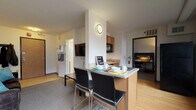 Located at 505 N Frances St in Madison, Wisconsin, this single-floor residential unit offers a functional and well-organized layout designed for comfortable living. The open-concept living and kitchen area serves as the heart of the unit, where a peninsula with seating for two provides a casual dining space. The kitchen is equipped with warm wood cabinetry, a stainless steel sink, a dishwasher, and an over-the-range microwave, offering practical everyday convenience. The living area features carpet flooring, a wall-mounted flat-screen TV, and a coat rack near the entry door, creating a welcoming atmosphere upon arrival. Two bedrooms are carpeted and provide a quiet retreat, each furnished with a bed and storage. One bedroom includes a dedicated study desk with built-in shelving, ideal for academic or professional use, while the other features a dresser and closet access with views to an adjacent area. The shared bathroom is appointed with a double-sink vanity featuring warm wood cabinetry, a white countertop, tile flooring, and a shower enclosure with a sliding glass door. Light wood-style flooring runs through the entry and kitchen areas, complementing the clean, neutral color palette found throughout the unit.
