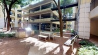 This outdoor courtyard is a well-appointed amenity space situated at the ground level of a multi-story residential complex. The area is defined by its open-air design, framed on multiple sides by the building's tiered balconies and colonnaded parking structure, creating a sheltered and semi-enclosed atmosphere.

At the heart of the courtyard stands a prominent built-in outdoor kitchen station, featuring a stainless steel gas grill set into a stone masonry surround with integrated storage — ideal for outdoor cooking and entertaining. Adjacent to the grill, a modern white dining table with matching benches and chairs provides a dedicated space for al fresco meals.

Mature trees anchor the space, offering natural shade across the red earth ground covering, which gives the courtyard a relaxed, organic character. Classic globe-style lamp posts are positioned throughout, ensuring the area is well-lit during evening hours. Additional seating benches are available around the perimeter for casual gathering.

The surrounding multi-level building façade, finished in warm neutral tones with wrought iron balcony railings, adds architectural continuity and a sense of enclosure. The integrated covered parking below the building levels is visible along the edges of the courtyard, underscoring the practical design of the complex while maintaining an inviting outdoor social environment.
