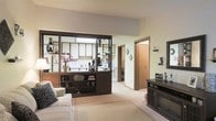 This single-floor residence offers a thoughtfully arranged layout that balances openness with defined living zones. The living area is anchored by plush, neutral-toned carpeting and furnished with a comfortable sofa, creating a welcoming atmosphere. A standout architectural feature is the dark-framed, open shelving room divider that elegantly separates the living space from the kitchen while maintaining a visual connection between the two areas. The divider incorporates built-in shelving for display and storage, adding both function and character to the space.

The kitchen, visible through the divider, benefits from a skylight that draws in natural light, brightening the workspace and enhancing the airy feel of the interior. Cabinetry in the kitchen is finished in a clean, light tone that complements the overall neutral palette of the home.

Warm wood-toned door frames and trim accents run throughout the space, adding a cohesive design element that ties the rooms together. A dark wood media console along the living room wall provides additional storage and display space. The corridor visible from the living area suggests further rooms beyond, completing this well-organized and comfortably appointed single-level home.