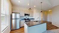 Located at 6650 Liberty Grand Blvd. in Powell, Ohio, this single-floor residence offers a thoughtfully designed layout with modern finishes throughout. The kitchen serves as a central highlight, showcasing dark granite countertops, white shaker-style cabinetry, and a large center island with a built-in sink. Stainless steel appliances — including a refrigerator, electric range, and over-the-range microwave — complement the space, while pendant lighting and recessed ceiling lights add warmth and functionality. Light wood-look flooring flows through the kitchen and main living corridor, creating a cohesive, open feel. Large windows throughout allow natural light to fill the space, with views of the surrounding community. The bedrooms are finished with plush carpeting and feature dedicated closets, including a walk-in closet adjacent to the primary bathroom. The bathroom includes a full tub-shower combination with clean white surroundings and wood-look flooring. A dedicated utility room houses a full-size washer and dryer alongside the water heater and HVAC system. The overall design balances practical functionality with clean, contemporary aesthetics, making this a well-appointed single-story residence.