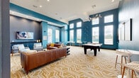 This single-floor community amenity space is a thoughtfully designed lounge and game room that combines social functionality with striking interior design. The room is defined by its soaring ceilings and bold teal-painted walls, creating a lively and energetic atmosphere throughout. Large floor-to-ceiling windows line the perimeter, flooding the space with generous natural light and offering views of the surrounding exterior grounds.

The patterned gold-and-white geometric carpet anchors the room and adds a layer of visual texture that ties together the eclectic furnishings. A full-size billiards table occupies a central position, making it a natural focal point for social gatherings, while a foosball table provides additional recreational options nearby. High-top bistro tables and chairs are positioned near the windows, offering bright, casual spots to sit and relax.

The lounge area features a pair of rich leather sofas and accent chairs arranged around a flat-screen TV, with a decorative feature wall showcasing an abstract gold-and-white wallcovering flanked by floating shelves displaying curated décor. Recessed lighting and a modern chandelier provide warm ambient illumination. A darkened accent nook with built-in cabinetry adds depth and dimension to the overall design. This space is ideal for socializing, recreation, and community gatherings.