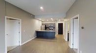 This two-bedroom, two-bathroom residence is thoughtfully designed across a single floor, offering a contemporary open-concept layout that seamlessly connects the living and kitchen areas. The kitchen serves as a focal point, featuring dark navy cabinetry paired with stainless steel appliances, including a refrigerator, range, and built-in microwave. A peninsula with a light-toned countertop extends into the living space, providing both additional prep space and casual seating opportunities. Recessed lighting throughout ensures consistent, even illumination across the open-plan area. The flooring features light wood-tone planks that run continuously through the main living space, lending warmth and visual cohesion. Neutral gray walls complement the modern cabinetry and trim details. The entryway is anchored by a dark front door, with a hallway leading to the two bedrooms and bathrooms. White trim and clean lines throughout the space contribute to a polished, move-in-ready aesthetic. The overall layout is efficient and well-organized, making the most of the available footprint while maintaining an open, airy feel.