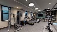 This contemporary fitness center offers a comprehensive workout environment designed to accommodate a wide range of exercise routines. The space is accessed through sleek, full-height glass-paneled doors framed in matte black, providing a transparent view into the facility and contributing to an open, professional aesthetic. The flooring throughout is a neutral-toned striped carpet, offering both durability and comfort underfoot.

The cardio zone features multiple treadmills equipped with individual digital screens, alongside an elliptical trainer, all arranged to maximize floor space. A dedicated strength and free weights area includes a multi-station cable machine, a flat weight bench, and an organized rack of dumbbells. Mirrors line portions of the walls, enhancing the sense of space and allowing users to monitor their form.

A standout functional training zone is anchored by built-in wood shelving units stocked with medicine balls and stability balls, while a row of kettlebells lines the base. A wall-mounted flat-screen television is set against a striking green subway tile accent wall, adding a bold design element to the space. Additional functional tools, including a BOSU balance trainer and a TAC plyo box, round out the equipment offerings.

Overhead, a combination of circular flush-mount lights and recessed spotlights provides bright, even illumination throughout. Windows along the perimeter allow for natural light, contributing to an energizing atmosphere well-suited for fitness activity.