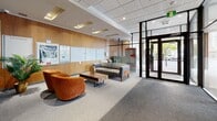 Located at 450 Rue Sherbrooke E in Montréal, this professionally designed lobby serves as an inviting gateway to the building. The space is defined by its modern aesthetic, combining a neutral grey carpet, a drop-panel ceiling with recessed lighting, and a striking dark tile accent wall that anchors the interior. Floor-to-ceiling glass walls and large glazed entrance doors flood the lobby with natural light while framing views of the surrounding streetscape, creating a seamless connection between the interior and the urban environment outside.

The seating area is thoughtfully furnished with a mid-century modern grey sofa and a wooden coffee table, complemented by a pair of bold orange accent chairs that add a warm pop of colour. Decorative elements, including a vibrant green tree artwork and potted plants, bring life and freshness to the space. A sleek console table with sculptural décor accents the dark feature wall.

Towards the building's interior corridor, a bank of resident mailboxes is neatly integrated into a warm wood-panelled wall, providing both function and visual warmth. A large framed mirror near the entrance enhances the sense of depth and space. The lobby's well-considered layout balances welcoming comfort with efficient circulation, making it an impressive and functional point of arrival.