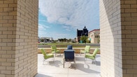 This single-level space features a well-appointed outdoor terrace that serves as its standout feature. Framed by light-colored brick walls on either side, the open-air patio creates a naturally sheltered seating area that opens dramatically toward a sweeping urban skyline. The terrace is finished with a smooth concrete surface and furnished with a curated arrangement of green-cushioned chairs and navy blue lounge seating, centered around a low coffee table — ideal for casual gathering or relaxation.

A low perimeter wall defines the boundary of the terrace while preserving unobstructed sightlines to the surrounding cityscape, which includes a mix of mid-rise commercial buildings, green spaces, and architectural landmarks. Natural light floods the space throughout the day, and the open sky above lends an expansive, airy quality to the setting.

The overall ambiance is calm and inviting, balancing urban energy with a comfortable outdoor retreat. The architectural framing of the brick columns adds structure and a sense of privacy without enclosing the space, making it a distinctive and functional area for enjoying the outdoors and taking in the surrounding environment.