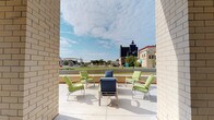 This single-level space features a well-appointed outdoor terrace that serves as its standout feature. Framed by light-colored brick walls on either side, the open-air patio creates a naturally sheltered seating area that opens dramatically toward a sweeping urban skyline. The terrace is finished with a smooth concrete surface and furnished with a curated arrangement of green-cushioned chairs and navy blue lounge seating, centered around a low coffee table — ideal for casual gathering or relaxation.

A low perimeter wall defines the boundary of the terrace while preserving unobstructed sightlines to the surrounding cityscape, which includes a mix of mid-rise commercial buildings, green spaces, and architectural landmarks. Natural light floods the space throughout the day, and the open sky above lends an expansive, airy quality to the setting.

The overall ambiance is calm and inviting, balancing urban energy with a comfortable outdoor retreat. The architectural framing of the brick columns adds structure and a sense of privacy without enclosing the space, making it a distinctive and functional area for enjoying the outdoors and taking in the surrounding environment.