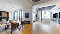 Located at 690 N Sherman St. in Denver, CO, this single-floor space offers a thoughtfully designed amenity suite with a contemporary aesthetic throughout. Light hardwood flooring runs continuously across the entire floor, lending warmth and cohesion to the various distinct zones within the space.

The entry area greets visitors with an elevator foyer accented by geometric tile wallcovering, opening directly into a casual lounge with mid-century-inspired seating. Floor-to-ceiling black-framed windows frame sweeping views of the Denver skyline, flooding the space with natural light. A dedicated billiards room features dramatic double-height ceilings with a dark-painted ceiling plane and a statement pendant fixture above a contemporary pool table.

The kitchen and dining area is anchored by a large white island beneath striking navy pendant lights, with dark cabinetry and a hex-tile backsplash housing a bar sink and beverage refrigerator. A formal lounge area nearby features a bold circular chandelier, built-in shelving, and a wall-mounted television set within a dark-paneled niche. A glass-partitioned conference room offers a professional meeting environment, while the outdoor terrace extends the usable area with panoramic city and mountain views.