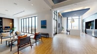 Located at 690 N Sherman St. in Denver, CO, this single-floor space offers a thoughtfully designed amenity suite with a contemporary aesthetic throughout. Light hardwood flooring runs continuously across the entire floor, lending warmth and cohesion to the various distinct zones within the space.

The entry area greets visitors with an elevator foyer accented by geometric tile wallcovering, opening directly into a casual lounge with mid-century-inspired seating. Floor-to-ceiling black-framed windows frame sweeping views of the Denver skyline, flooding the space with natural light. A dedicated billiards room features dramatic double-height ceilings with a dark-painted ceiling plane and a statement pendant fixture above a contemporary pool table.

The kitchen and dining area is anchored by a large white island beneath striking navy pendant lights, with dark cabinetry and a hex-tile backsplash housing a bar sink and beverage refrigerator. A formal lounge area nearby features a bold circular chandelier, built-in shelving, and a wall-mounted television set within a dark-paneled niche. A glass-partitioned conference room offers a professional meeting environment, while the outdoor terrace extends the usable area with panoramic city and mountain views.