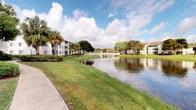 This single-story residence is part of a well-established multi-building community set against a picturesque Florida backdrop. The exterior environment is a standout feature, with the community centered around a calm, reflective pond surrounded by manicured green lawns, mature palm trees, and broad-canopied shade trees. The buildings are finished in light neutral tones with accented sliding doors and balconies, creating a cohesive coastal aesthetic that blends naturally into the tropical landscape.

A paved walkway runs alongside the waterfront, offering residents a pleasant path for leisurely strolls with unobstructed views of the pond and its mirror-like surface. A pergola-style structure near the water's edge adds an architectural accent to the outdoor amenity space, enhancing the community's resort-like ambiance. The grounds are meticulously maintained, with open green spaces providing a sense of openness and connection to nature.

The surrounding environment is characterized by abundant natural light, sweeping sky views, and the calming presence of water throughout the community. The overall atmosphere is peaceful and scenic, reflecting a lifestyle oriented around outdoor living and natural beauty typical of Florida residential communities.