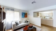This single-floor space showcases a thoughtfully designed open layout that seamlessly connects the living area to the kitchen. The living room is bright and airy, benefiting from a large window dressed with vertical blinds that allow generous natural light to filter in. White walls provide a clean, neutral backdrop, accentuated by a bold, colorful abstract artwork that adds a vibrant focal point to the room. The space accommodates a contemporary white sofa adorned with an assortment of decorative pillows, complemented by a set of uniquely shaped geometric accent tables in red and dark tones atop a plush area rug. Warm hardwood-style flooring runs throughout, adding texture and continuity between rooms. A pass-through opening in the wall connects the living area to the kitchen, which features light wood cabinetry, white countertops, and recessed under-cabinet lighting. The kitchen maintains a clean, functional aesthetic with ample cabinet storage. Overall, the space offers a cohesive, modern feel with a warm and inviting ambiance suitable for both everyday living and entertaining.