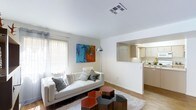 This single-floor space showcases a thoughtfully designed open layout that seamlessly connects the living area to the kitchen. The living room is bright and airy, benefiting from a large window dressed with vertical blinds that allow generous natural light to filter in. White walls provide a clean, neutral backdrop, accentuated by a bold, colorful abstract artwork that adds a vibrant focal point to the room. The space accommodates a contemporary white sofa adorned with an assortment of decorative pillows, complemented by a set of uniquely shaped geometric accent tables in red and dark tones atop a plush area rug. Warm hardwood-style flooring runs throughout, adding texture and continuity between rooms. A pass-through opening in the wall connects the living area to the kitchen, which features light wood cabinetry, white countertops, and recessed under-cabinet lighting. The kitchen maintains a clean, functional aesthetic with ample cabinet storage. Overall, the space offers a cohesive, modern feel with a warm and inviting ambiance suitable for both everyday living and entertaining.