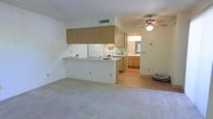 This single-story space presents an open-concept layout that integrates the living area and kitchen into one cohesive zone. The kitchen is outfitted with light wood cabinetry—including an overhead cabinet bank and lower storage units—alongside white appliances such as a refrigerator and range. A peninsula counter extends into the living space, offering a functional divide between the two areas while maintaining an open feel. The living area features neutral wall-to-wall carpeting, contributing to a clean, understated aesthetic. A ceiling fan with an integrated light fixture is mounted overhead, providing both air circulation and ambient lighting. A hallway visible beyond the kitchen leads to additional rooms, where a vanity mirror and cabinetry suggest a bathroom or dressing area with tile or hard flooring. The textured ceiling and light-toned walls throughout lend the space a bright, airy character. HVAC ventilation is integrated into the ceiling, and the overall layout is practical and straightforward, making efficient use of the available floor plan.