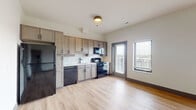 This single-floor, two-bedroom, two-bathroom residence in Louisville's Belma neighborhood offers a clean, contemporary design with consistent light wood-tone flooring throughout the main living areas. The open-concept kitchen is equipped with black appliances—including a refrigerator, gas range, microwave, and dishwasher—complemented by warm taupe cabinetry and a herringbone-patterned tile backsplash. A door and multiple windows in the living and kitchen area allow natural light to fill the space while providing outdoor views. The living area is generously proportioned, with a dedicated nook housing a stacked washer and dryer for in-unit convenience. A well-lit hallway connects the two bedrooms, each featuring hardwood-style flooring and reach-in closets with wire shelving. Both bathrooms feature marble-look tile flooring and matte black fixtures, lending a polished, modern aesthetic. One bathroom includes a soaking tub with a tiled surround. The overall design maintains a neutral, contemporary palette throughout, offering a clean and functional living environment.