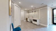 Located at 5430 Cane Ridge Rd in Nashville, TN, this single-floor commercial suite offers a thoughtfully designed layout comprising a hallway, a private office, and a bathroom. The main area greets visitors with large-format tile flooring, recessed lighting, and a neutral gray-and-white color palette that lends a clean, professional atmosphere throughout. A built-in kitchenette station anchors one wall, complete with a countertop, sink, under-counter appliances including a microwave and dishwasher, and a textured accent wall illuminated by a row of sleek pendant lights — adding a polished, modern touch. Crown molding and chair-rail detailing run along the walls, contributing to a refined finish. A glass-paneled entry door allows natural light to filter in from the exterior, brightening the main area. The private office is carpeted with a patterned tile-style carpet and features recessed lighting, offering a quiet, focused workspace. The bathroom is ADA-accessible, equipped with grab bars, a wall-mounted paper dispenser, and tile flooring with a floor drain, meeting functional commercial standards. Together, these spaces create a compact yet fully equipped professional environment suitable for a variety of business uses.