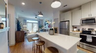 This thoughtfully designed single-floor residence offers a seamless open-concept layout connecting the kitchen, dining, and living areas. The kitchen is a standout feature, showcasing light gray shaker-style cabinetry, quartz countertops, a large center island with bar seating, and a full suite of stainless steel appliances including a refrigerator, range, microwave, and dishwasher. Track and pendant lighting illuminate the space, complemented by warm wood-look flooring that flows throughout the main living areas. The living room is bright and welcoming, anchored by a ceiling fan and a set of full-length French doors that flood the space with natural light and provide outdoor access. The primary bedroom is generously sized, offering plush carpet, large windows with blinds and drape panels, and direct access to an en-suite bathroom featuring granite countertops and stylish gray cabinetry. A second bedroom also benefits from ample natural light and comfortable carpeting. The bathroom includes overhead cabinetry for storage, granite countertops, and wood-look flooring consistent with the rest of the home. A hallway connects the rooms efficiently, with a walk-in closet visible from the bathroom. Clean white walls, contemporary fixtures, and cohesive finishes throughout give this residence a modern yet inviting ambiance.