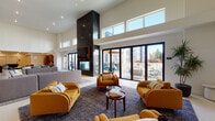 This single-floor amenity space is designed with a modern aesthetic, combining generous proportions, soaring double-height ceilings, and an abundance of natural light. The main lounge area serves as the centerpiece, anchored by a striking floor-to-ceiling dark-clad fireplace that commands attention. Plush mustard-yellow sofas and armchairs are arranged around a central coffee table atop a large area rug, creating an inviting gathering space. Expansive black-framed sliding glass doors open directly onto an outdoor terrace, flooding the interior with daylight and providing seamless indoor-outdoor connectivity. Clerestory windows positioned high on the walls further enhance the bright, airy atmosphere.

Adjacent to the lounge, a spacious dining area features a long dark communal table surrounded by modern teal chairs, complemented by curated décor including a circular gold mirror and a teal sideboard. The fully equipped kitchen offers warm wood cabinetry, stainless steel appliances, white quartz countertops, a large island with bar seating, and pendant lighting overhead. A dedicated lounge nook features a wall-mounted oversized Scrabble board, adding a playful, social element. Throughout, light-toned tile flooring, tall ceilings, and carefully selected furnishings contribute to a cohesive, welcoming communal environment.