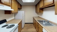 This single-floor residence offers a well-defined layout with distinct living zones connected by an open passage. The spacious living room is finished with neutral-toned carpet, a textured ceiling, baseboard heating, and a wall-mounted air conditioning unit. A large bank of windows along one wall allows generous natural light to filter in, providing views of the surrounding exterior.

An open architectural passage connects the living room to a dedicated dining area, distinguished by rich hardwood-style flooring, a ceiling fan with light fixture, and a dramatic full brick accent wall that serves as the standout design feature of the space. This area transitions seamlessly into the galley-style kitchen, which features warm wood cabinetry, light stone-finish countertops, a skylight overhead, a double sink, and white appliances including a range and refrigerator.

The bedrooms are carpeted and finished with neutral walls, textured ceilings, and individual windows with blinds, each equipped with baseboard heating. The bathroom area includes a wide vanity with white cabinetry, a large mirror, and dual overhead lighting fixtures, providing a practical and well-lit space.