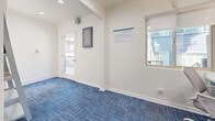 Located at 2401 S Hacienda Blvd in Hacienda Heights, CA, this single-floor business center offers a well-appointed and functional space designed for both work and relaxation. The main area is finished with vibrant blue patterned carpet tiles that add a modern, energetic feel against crisp white walls. A bold teal accent wall anchors the workstation zone, where a long white desk accommodates two computer monitors, complemented by task lamps and ergonomic chairs — creating an efficient and comfortable workspace. Natural light filters in through a glass-paneled exterior door and a side window, offering pleasant views of the surrounding outdoor area. A cozy reading nook is tucked alongside a lean ladder-style bookshelf stocked with an eclectic selection of books, paired with a plush blue armchair — perfect for quiet leisure. Wall-mounted decor, including a playful "The Book Was Better" canvas print, adds personality to the space. Recessed ceiling lights provide consistent illumination throughout. A utility room rounds out the layout, supporting the operational needs of the center. Overall, this space strikes a balance between a productive work environment and an inviting retreat for reading and relaxation.